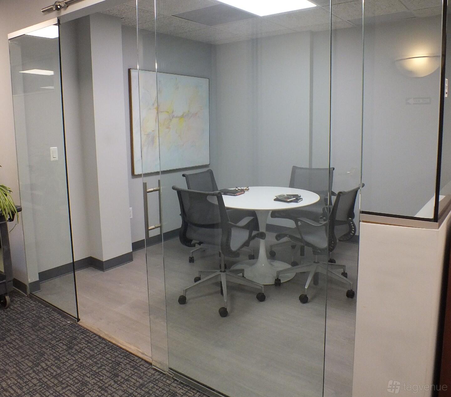A meeting room with a round white table, four mesh chairs, and abstract wall art at Office Suites of Darien.