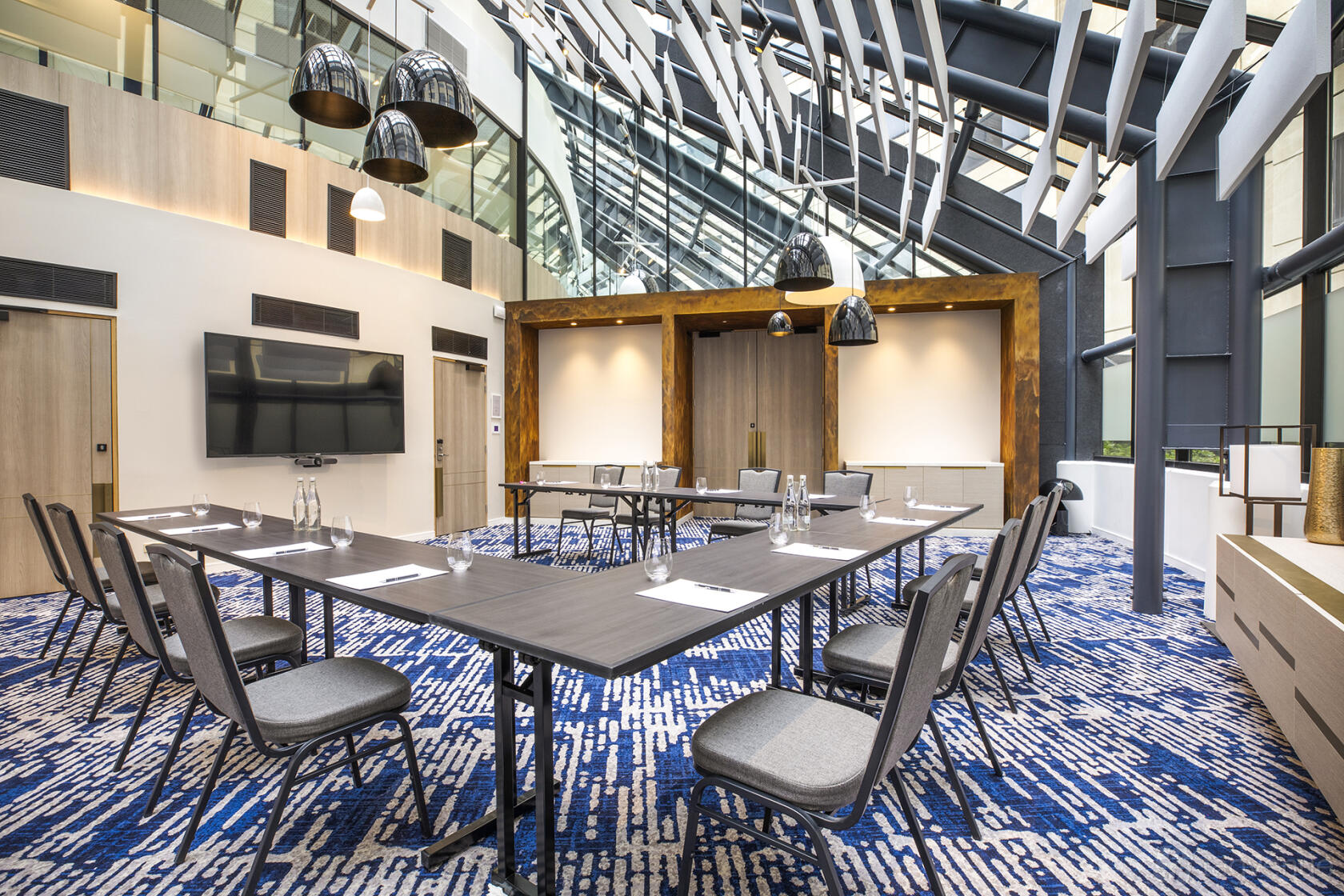 A meeting room with a glass ceiling, patterned carpet, pendant lighting, and U-shaped tables at Pearl Riverfront.