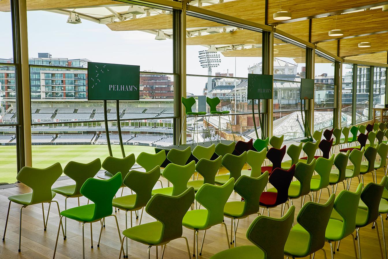 An event venue with floor-to-ceiling windows, colorful chairs, and stadium views at Lord's Cricket Ground.