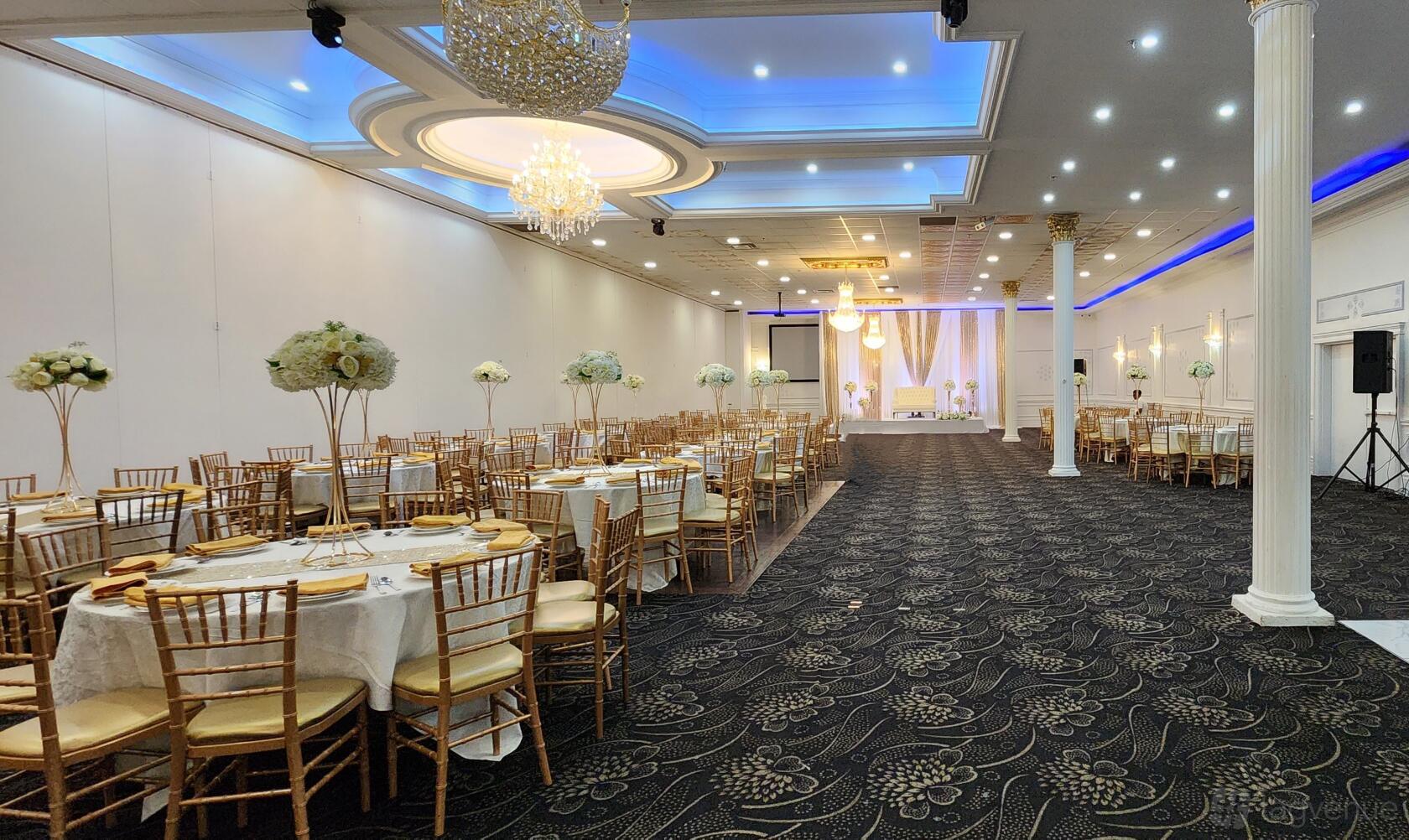 A banquet hall with gold chiavari chairs, round tables with white linens, chandeliers, and uplighting at Royal King Event Centre.