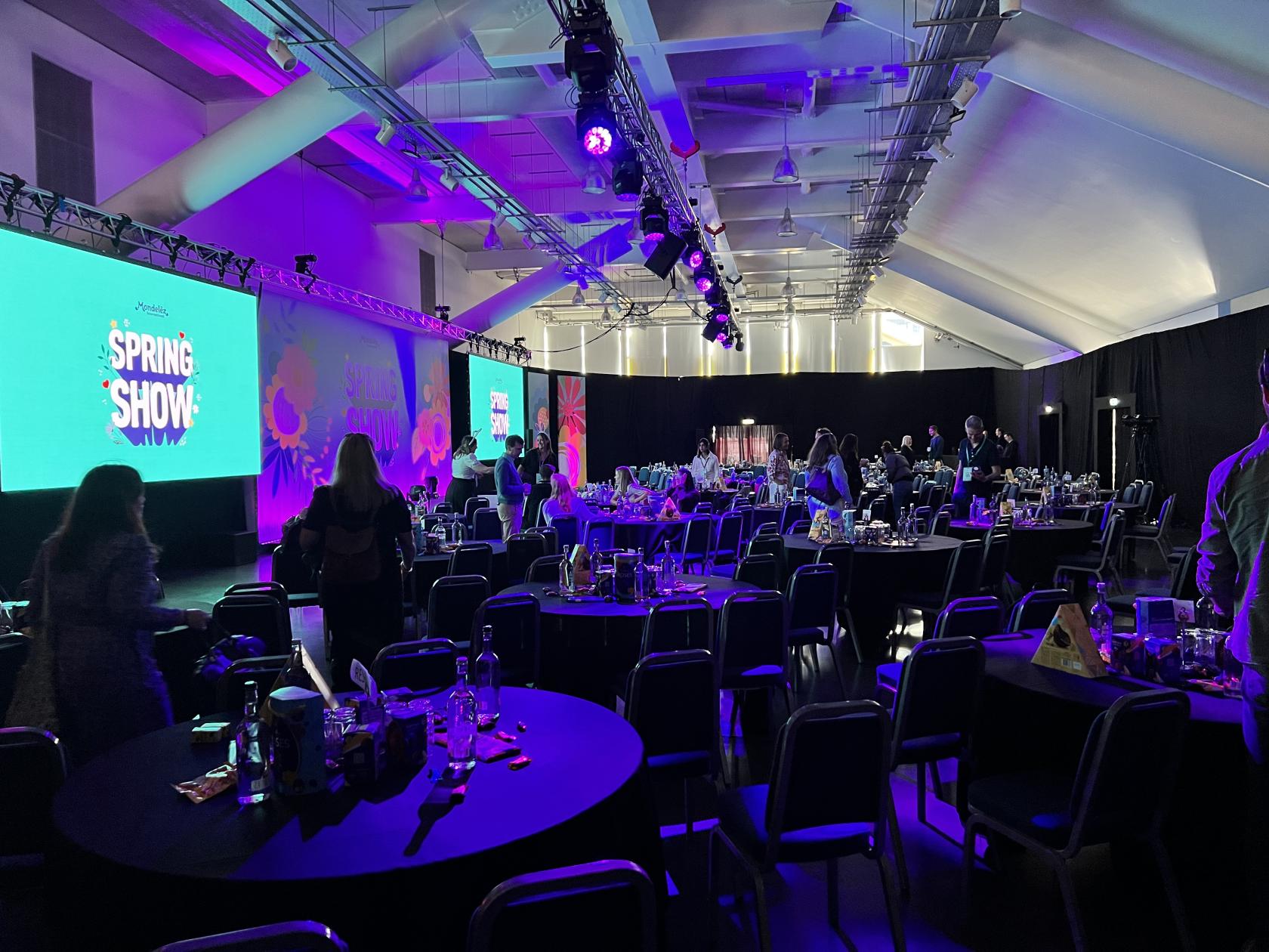 An event venue with round tables, chairs, large projection screens, and purple stage lighting at Newbury Racecourse.
