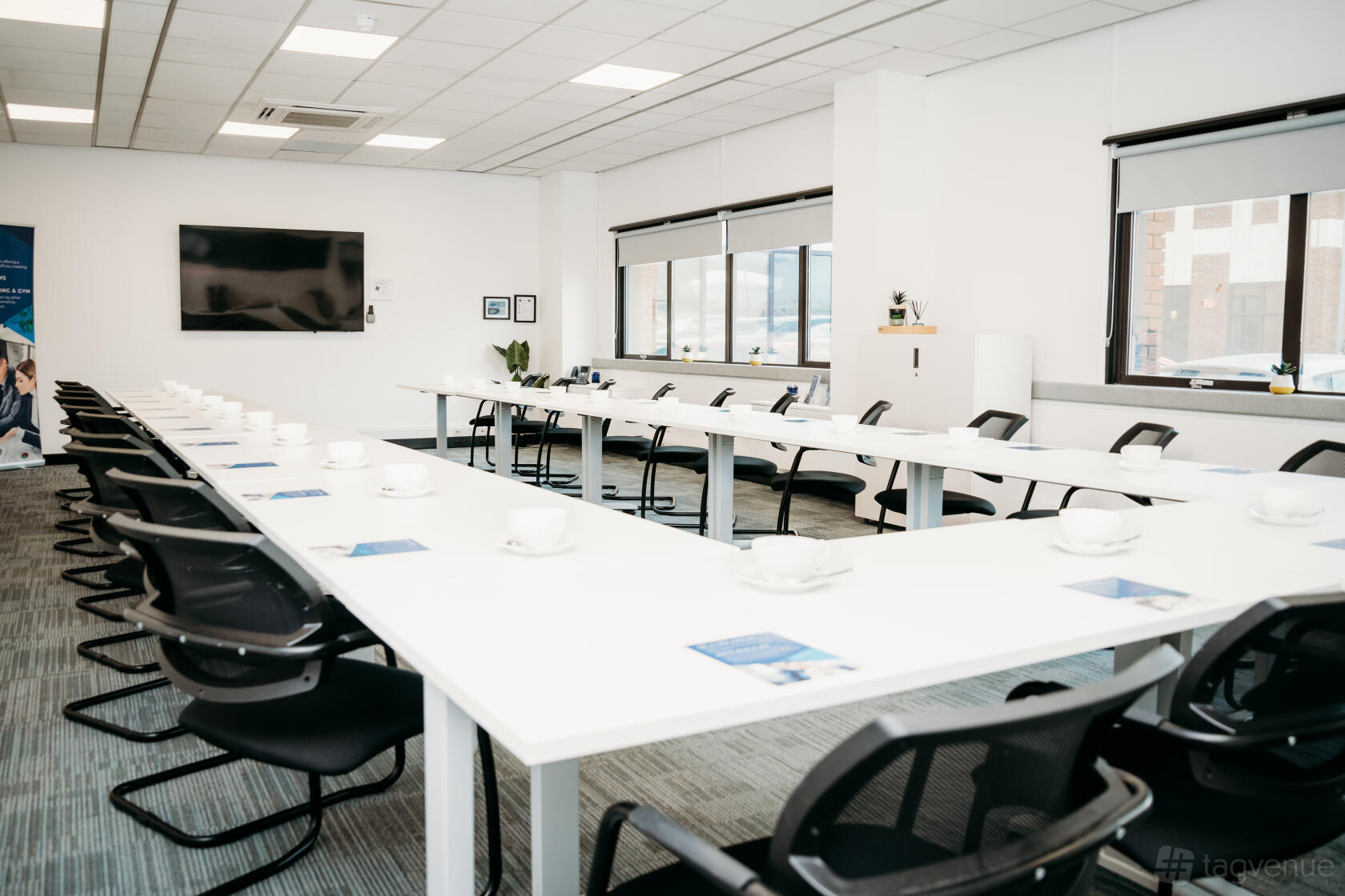 A meeting centre with U-shaped white tables, black chairs, large windows, and a wall-mounted TV at BusinessLodge Stoke.