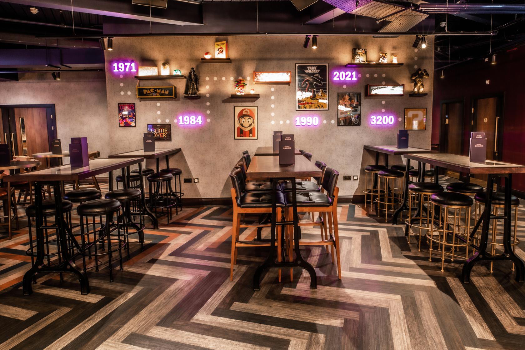 An event space with neon signage, framed wall art, herringbone wood flooring, and high tables at Puttshack Watford.