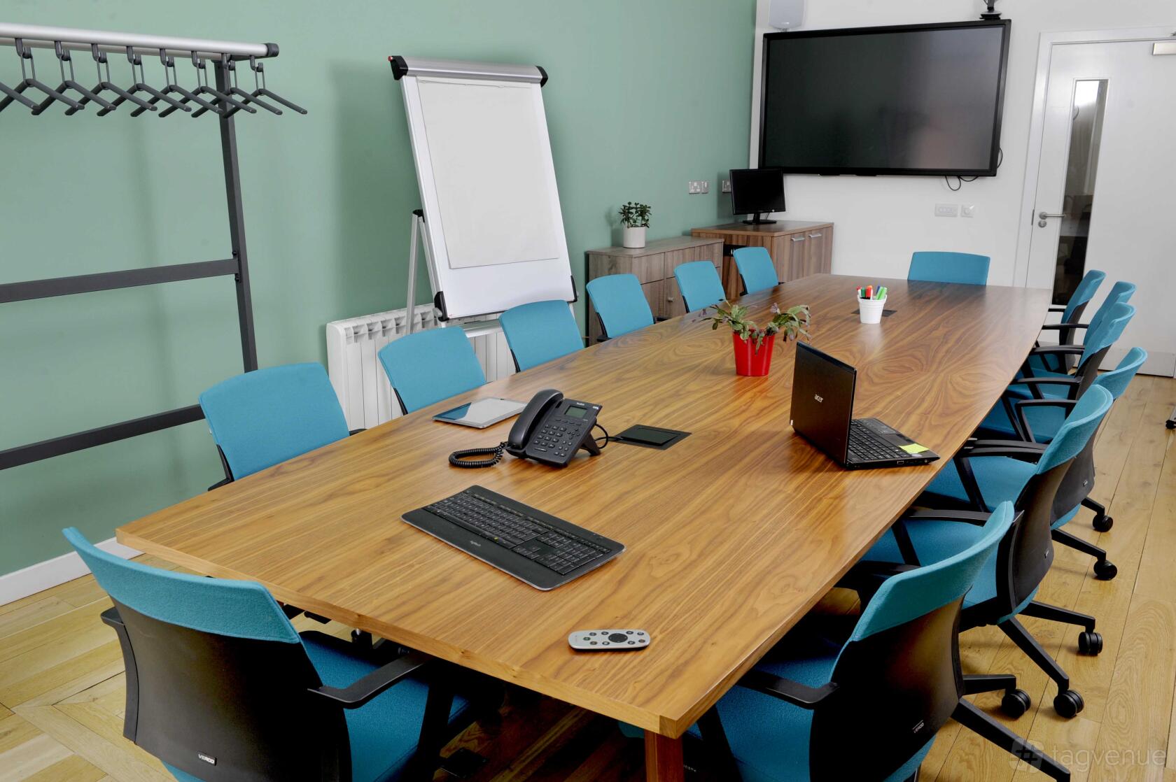 A boardroom with a long wooden table, turquoise chairs, flip chart, and wall-mounted screen at The Melting Pot.