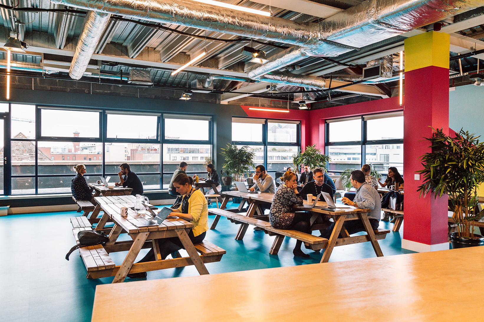 An event space with long wooden picnic tables, large windows, and exposed ductwork at Huckletree D2