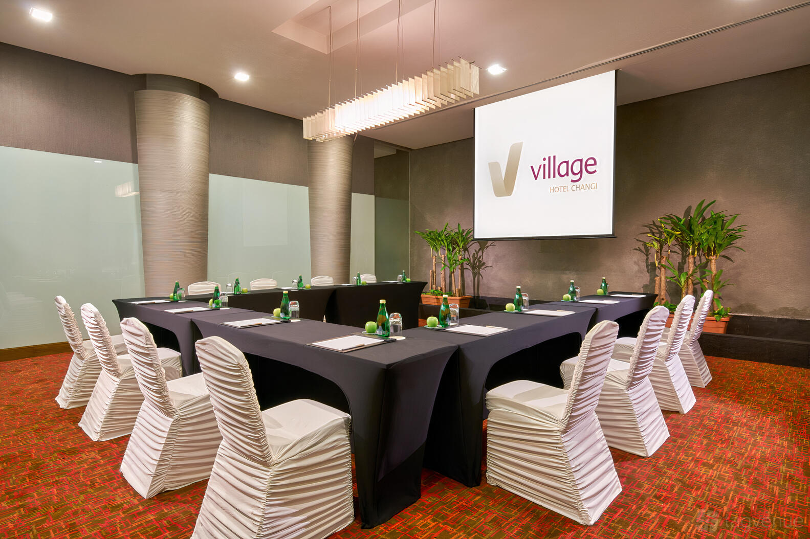 A meeting room with U-shaped tables, white chair covers, a projector screen, and potted plants at Village Hotel Changi.