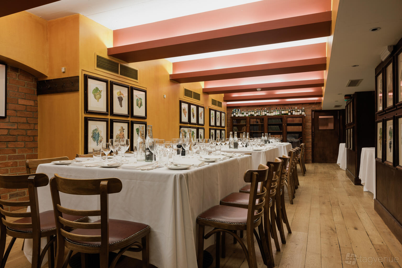 A dining room with long tables covered in white linens, wooden chairs, framed artwork, and a wall of wine bottles at Bleeding Heart.