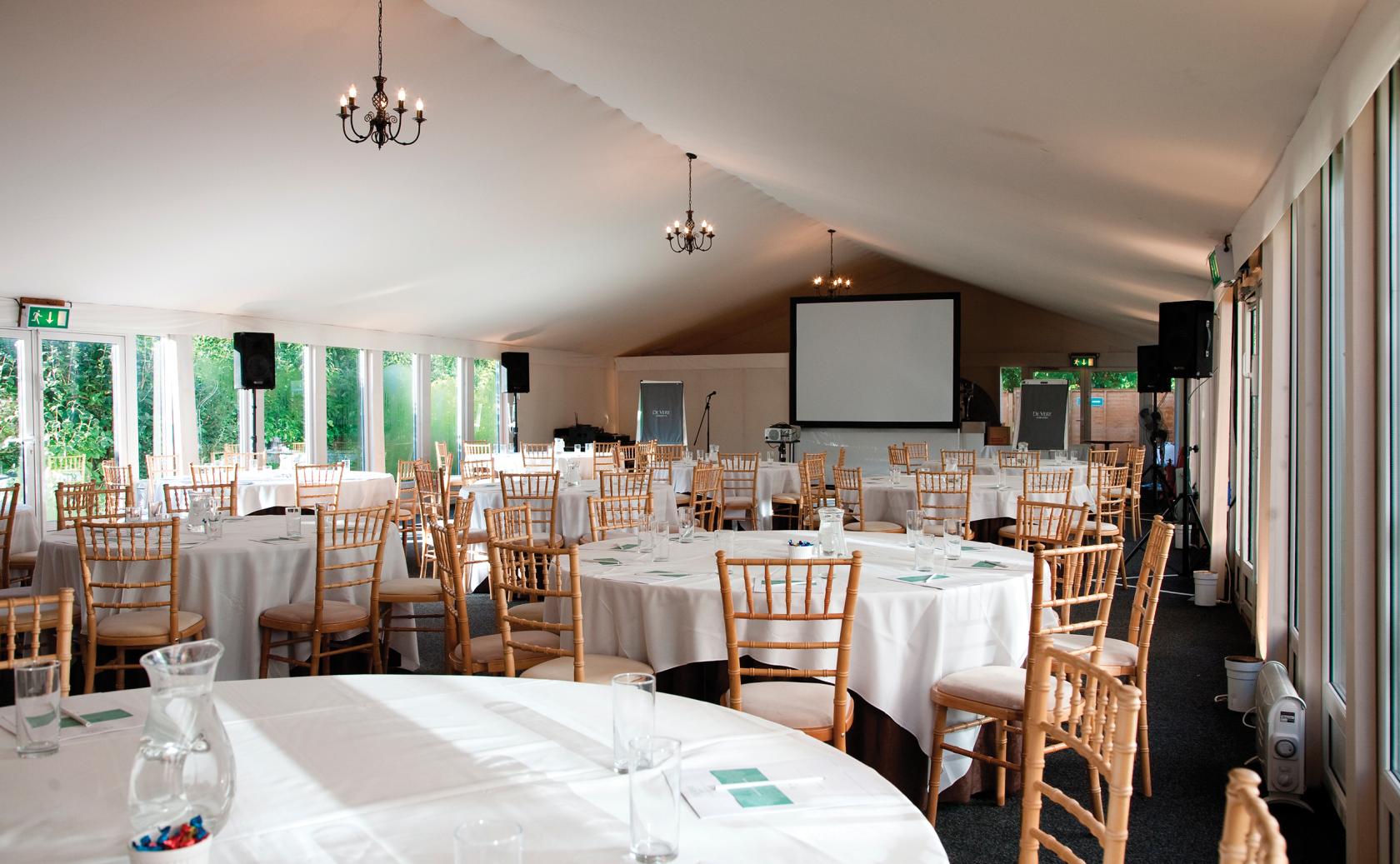 A terrace marquee with round tables, wooden chairs, chandeliers, and large windows at Hunton Park Hotel.