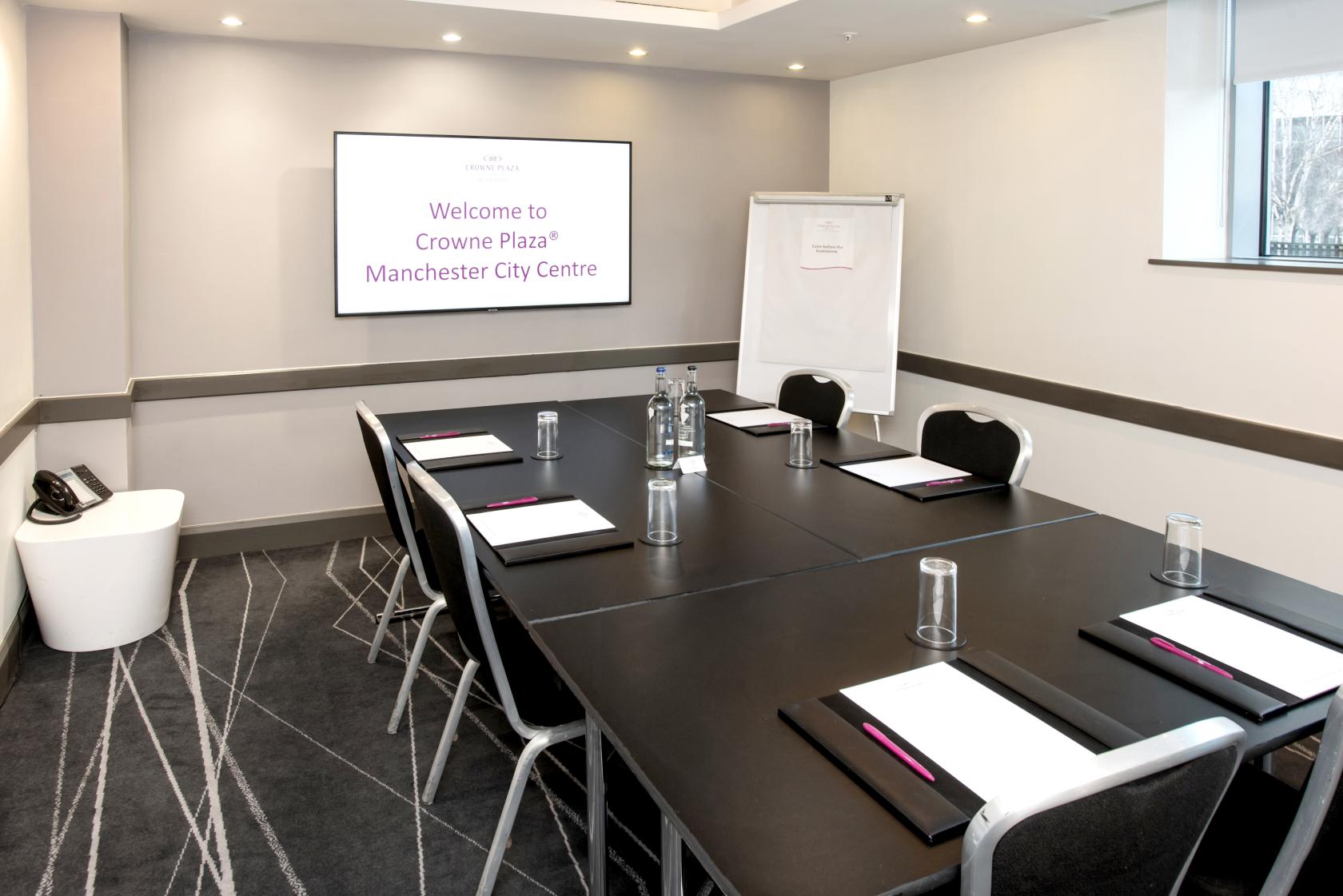 A boardroom with a rectangular table, black chairs, notepads, glass tumblers, and a wall-mounted screen at Crowne Plaza Manchester City Centre.