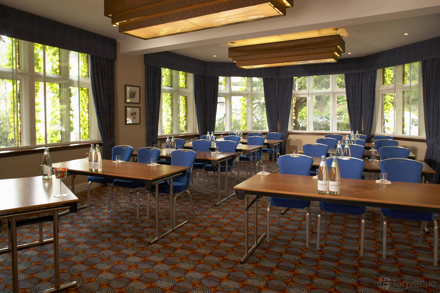 A function room with large windows, blue chairs, and conference tables set with bottled water at Abbey Hotel Worcester Suite.