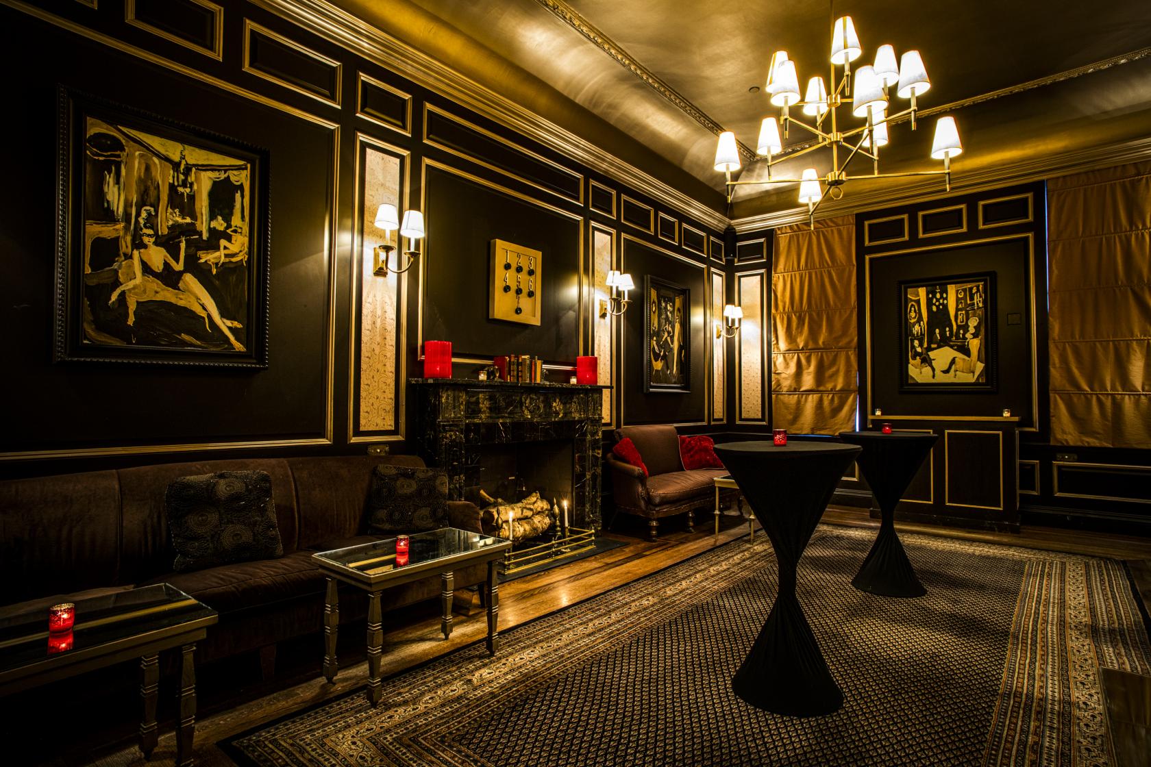 A function room with dark paneled walls, framed art, a decorative fireplace, and cocktail tables at The Raines Law Room at The William.