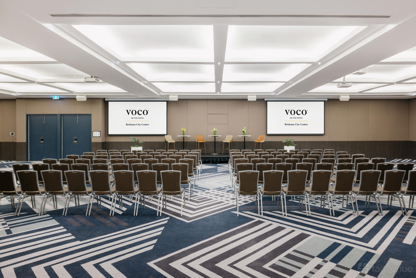 A hotel hall with rows of chairs, geometric-patterned carpet, and dual projection screens at Voco Hotel Brisbane City Centre.