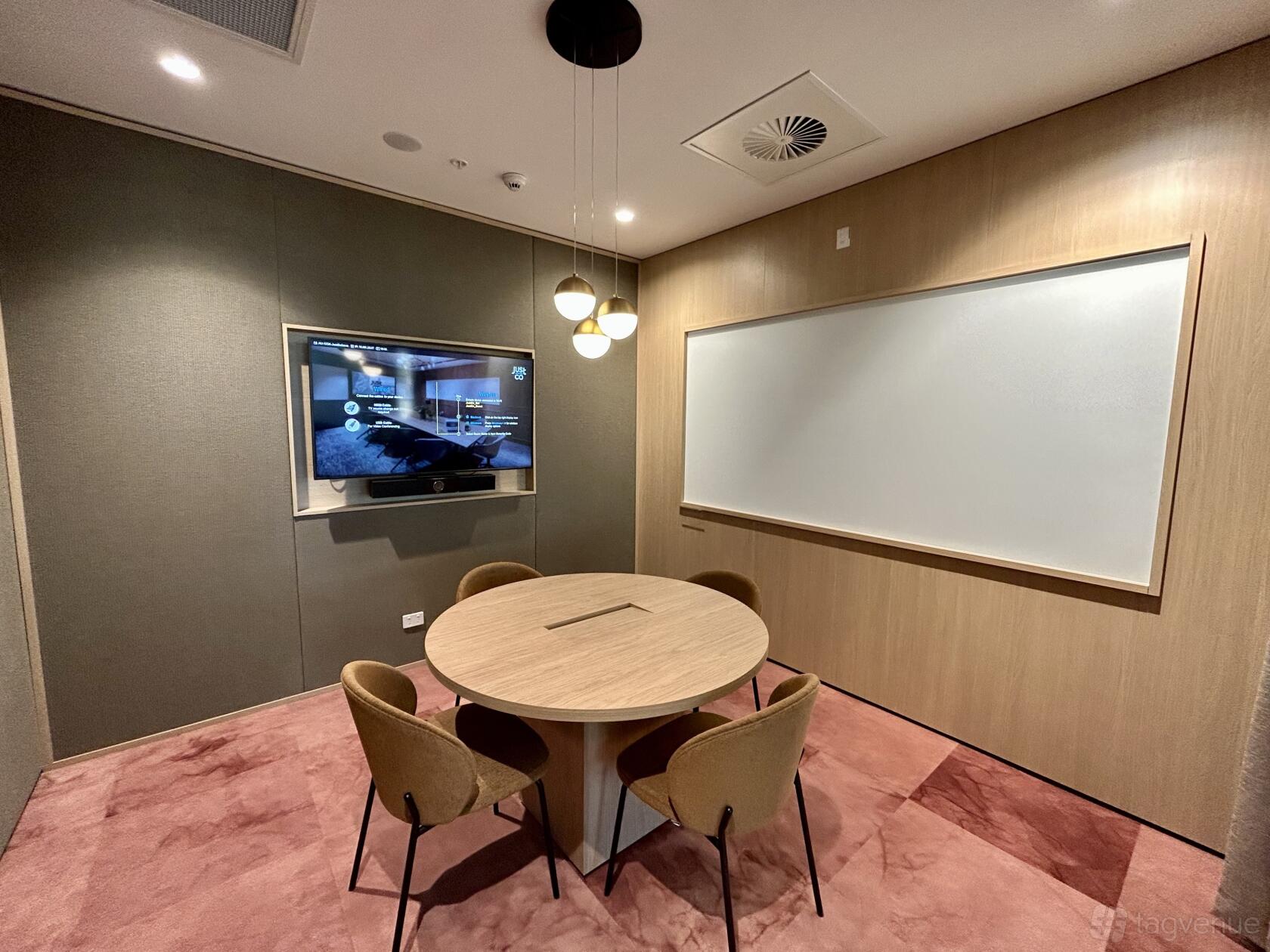 A meeting room with a round wooden table, four chairs, wall-mounted screen, and large whiteboard at JustCo at King Street.