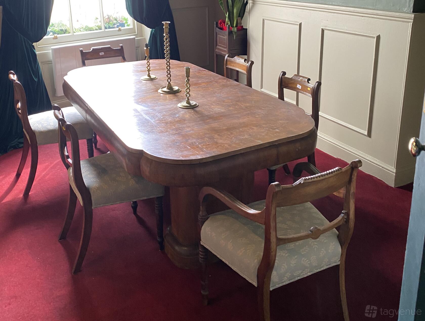 A dining room with a polished wooden table, six chairs, brass candlesticks, and red carpet at Brydges Place Club.