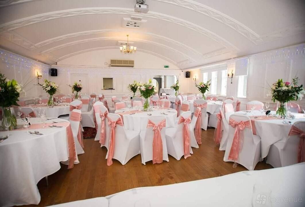 A function room with round tables draped in white linens, pink chair sashes, and floral centerpieces at Rose & Crown.