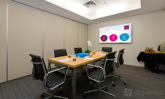 A meeting room with a wooden conference table, black leather chairs, and a wall-mounted screen at Sage Hotel Wollongong.