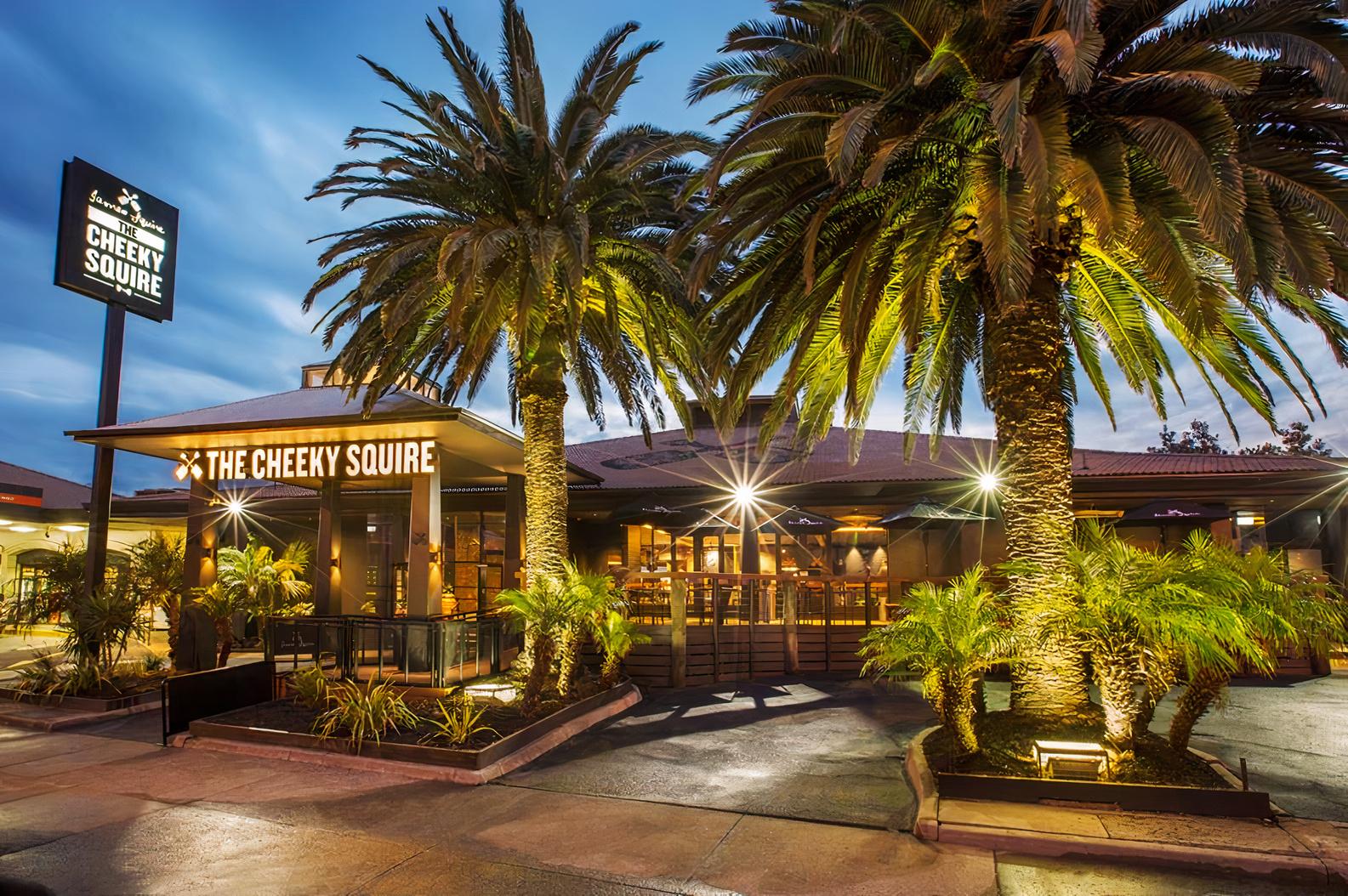 A bar with a large entrance patio, neon signage, and tall palm trees at The Cheeky Squire.