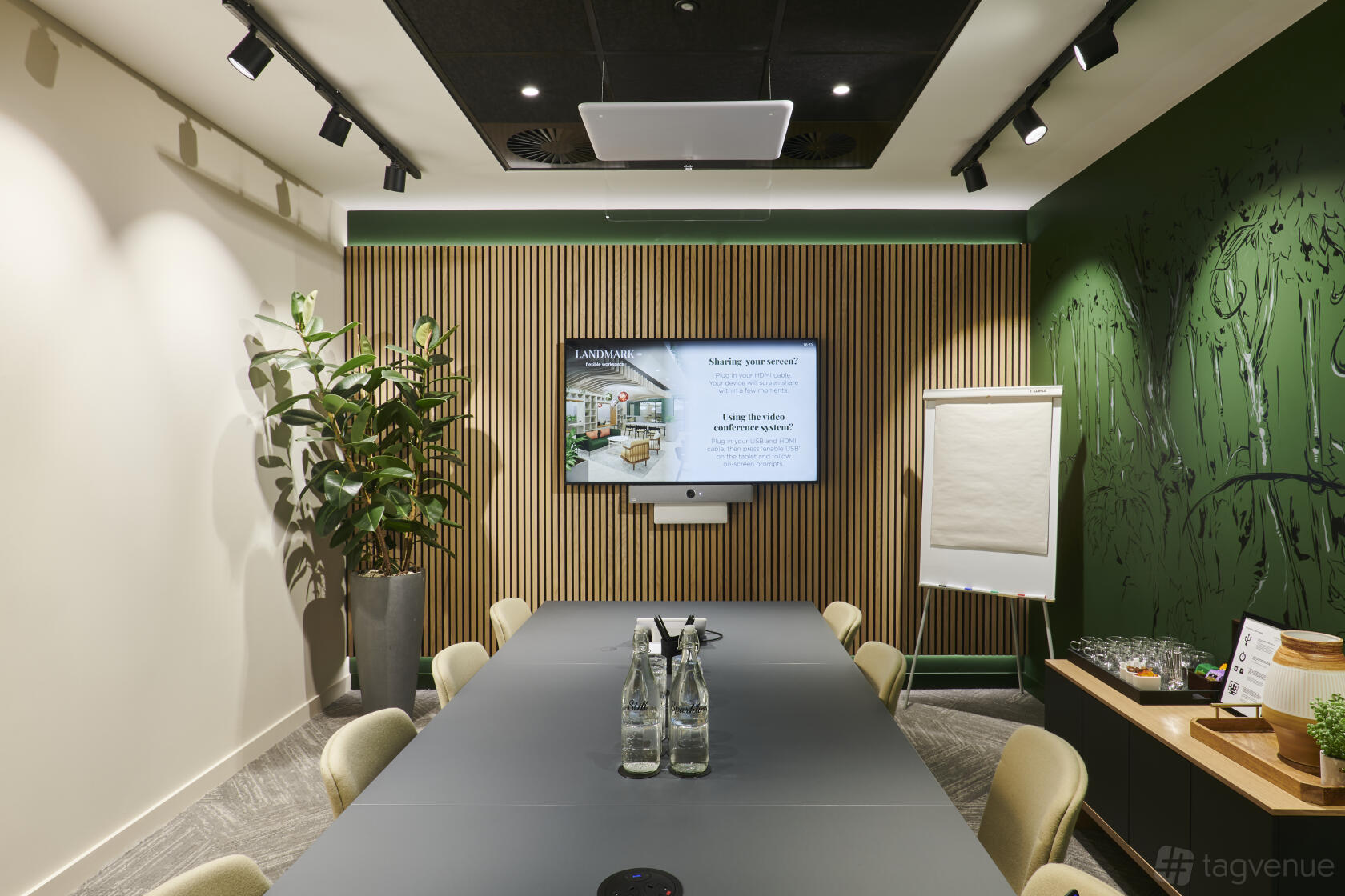 A meeting room with a long conference table, wall-mounted screen, flip chart, and green accent wall at Landmark: Orchard Place, Victoria, London.