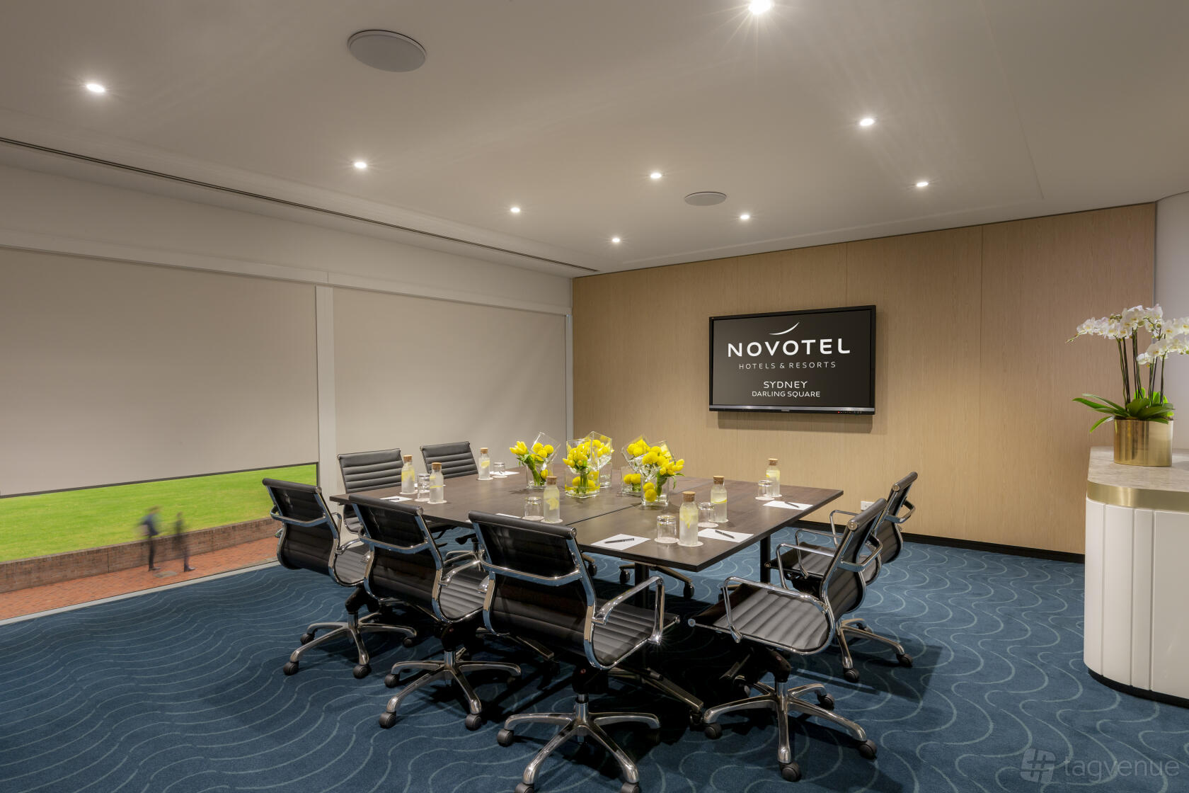 A hotel meeting room with a long table, black office chairs, large screen, and yellow floral centerpieces at Novotel Sydney Darling Square.