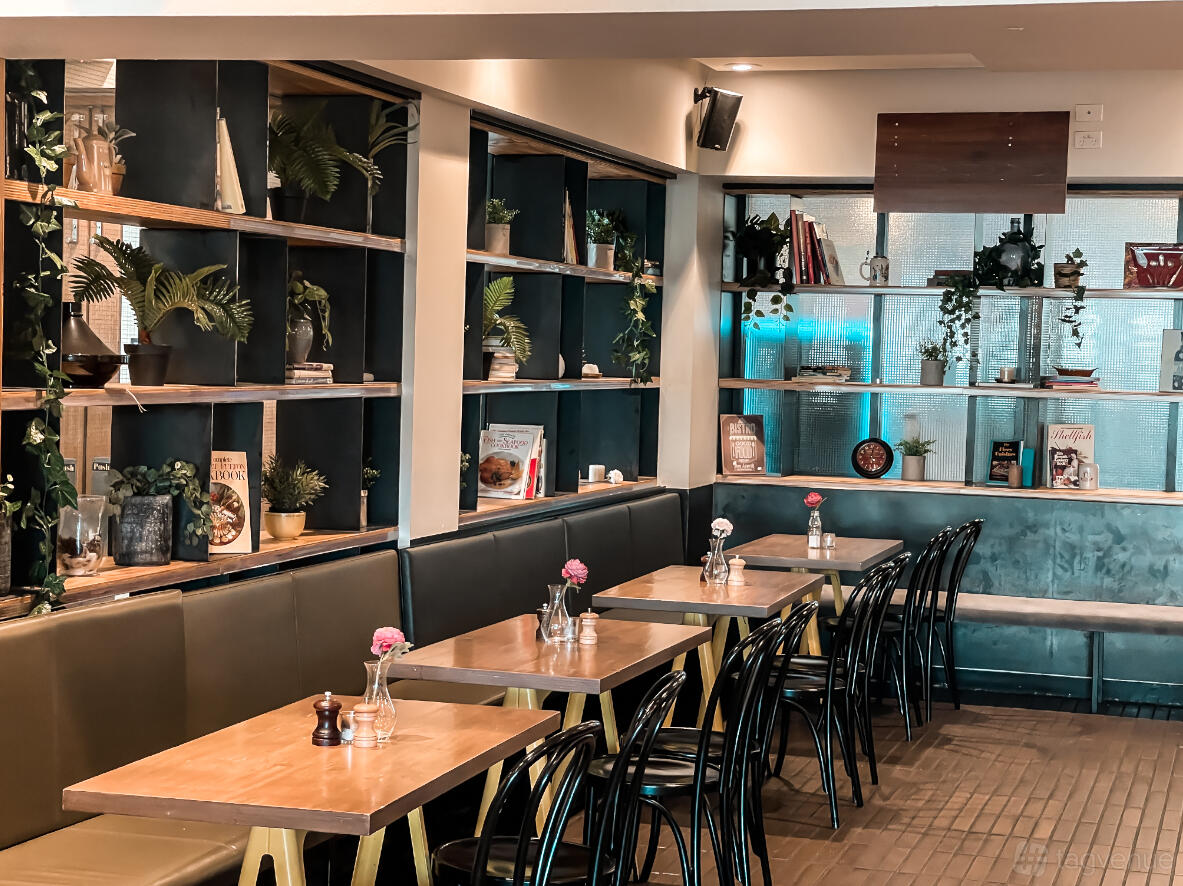 A pub interior with wooden tables, black chairs, built-in shelving with books and plants at Grosvenor Hotel.