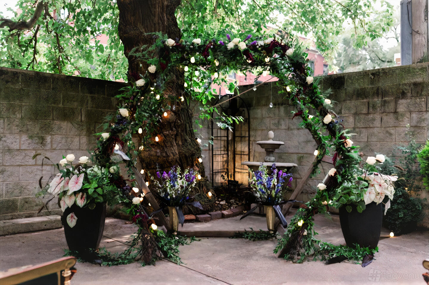 An outdoor event space with a floral arch, string lights, and stone fountain in the Courtyard at Colvin House.