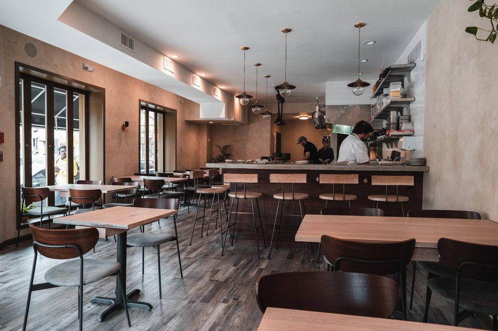 A restaurant with wooden tables, dark chairs, pendant lighting, and an open kitchen at River Twice.