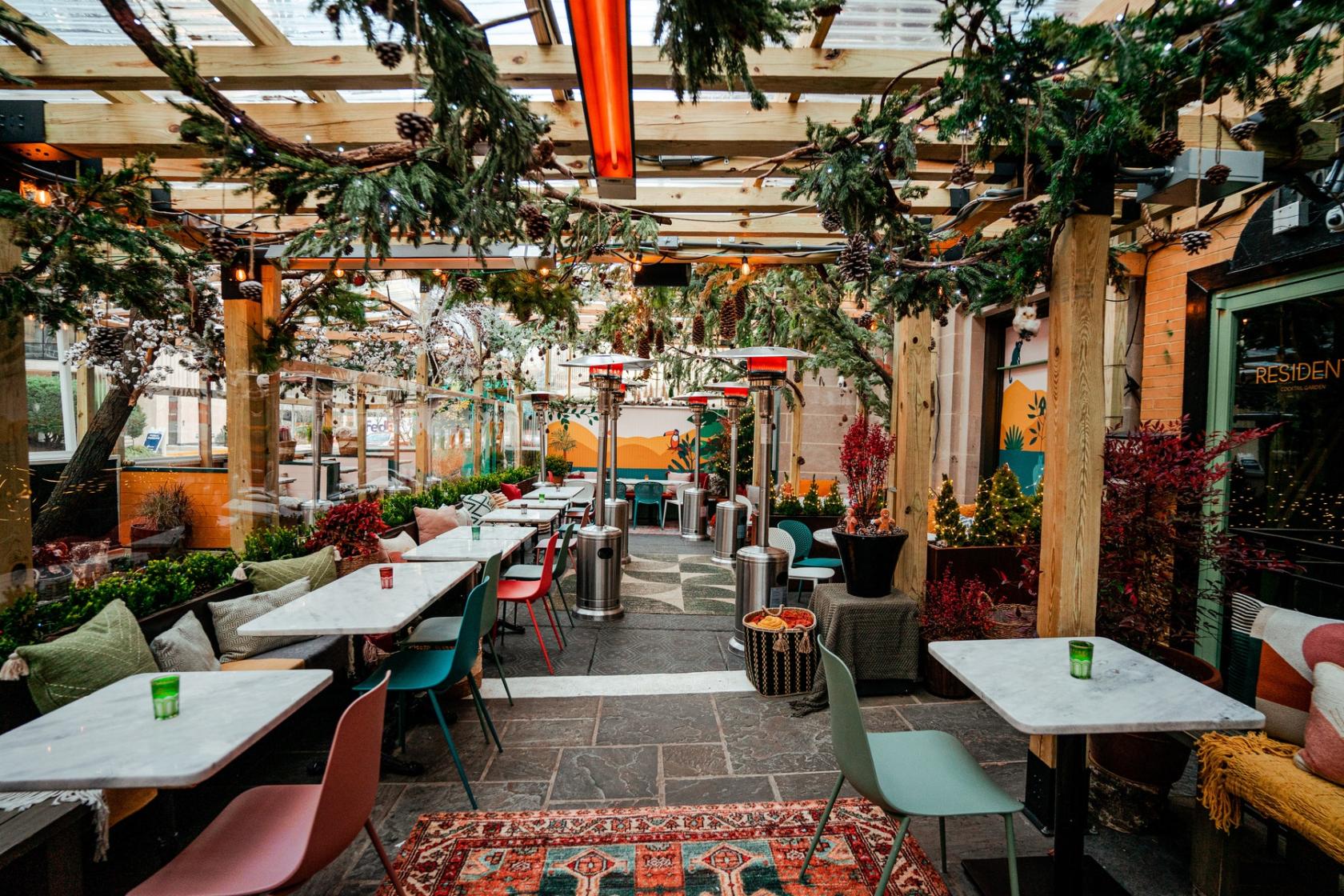 A restaurant patio with marble tables, colorful chairs, potted plants, and overhead greenery at Residents Cafe & Bar.