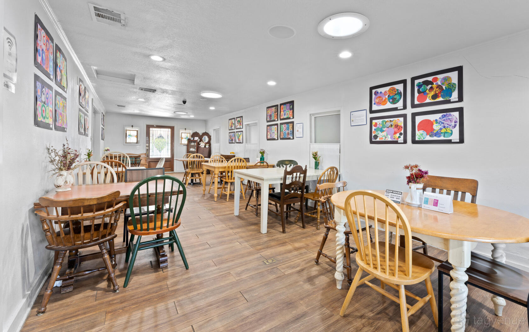 A cafe with wooden tables and mismatched chairs, framed artwork on white walls, and wood flooring at Brentwood Social House.