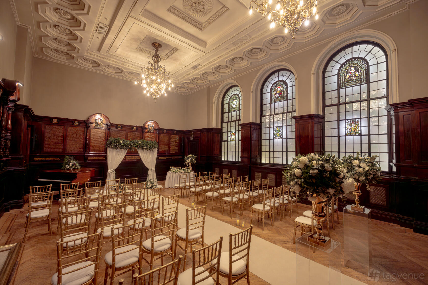 A function room with stained glass arched windows, chandeliers, and arranged gold chairs at Trades Hall of Glasgow.