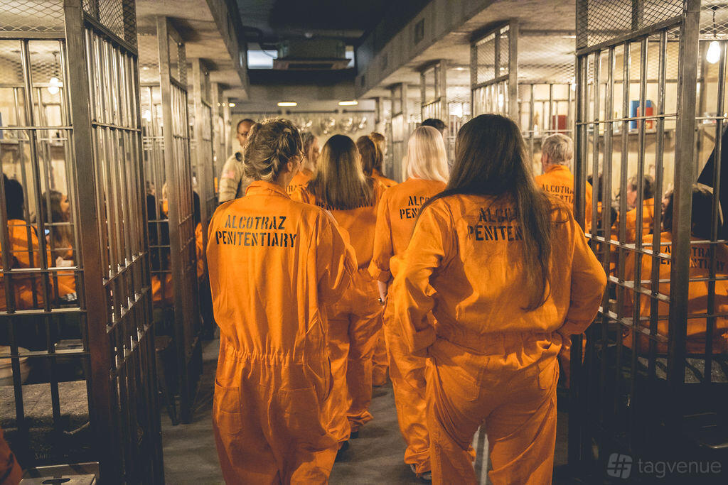 A cocktail bar with guests in orange prison jumpsuits walking between metal cell bars at Alcotraz London.