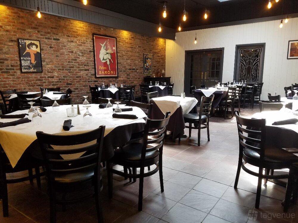 A bar with exposed brick walls, framed art, hanging bulbs, and round tables with white tablecloths at Bayonne Patio Bar & Grille.