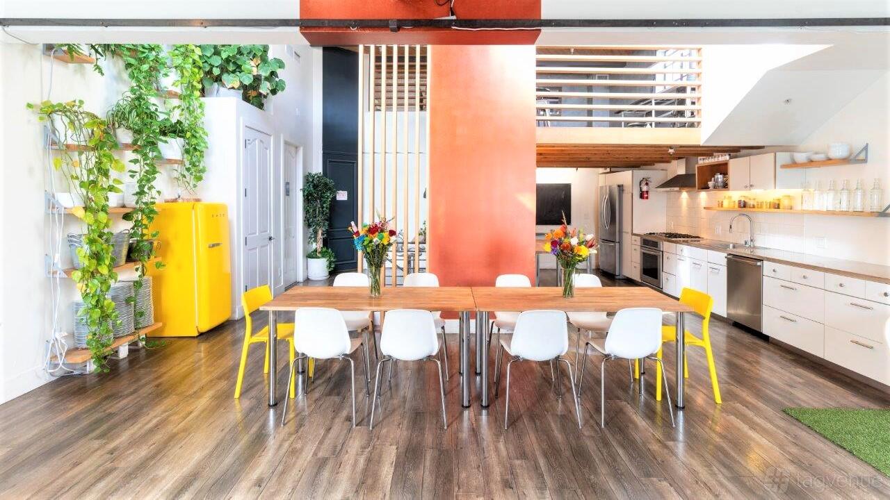 A loft event space with wood flooring, open kitchen, tall plants, and a long table with white chairs at Boutique Event & Meeting Loft.