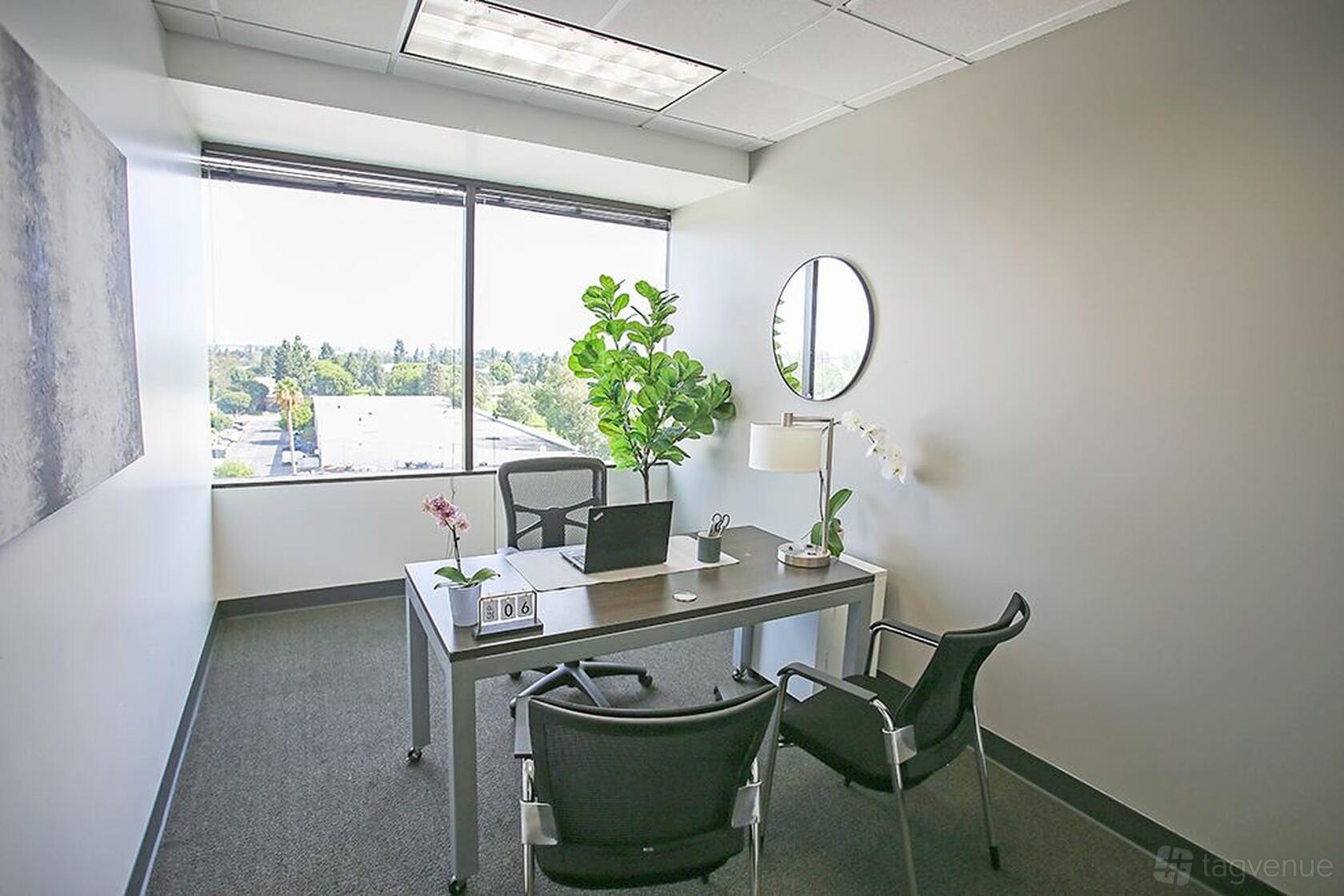 A meeting room with a large window, potted plants, and a desk with two chairs at Premier Workspaces - Cerritos.