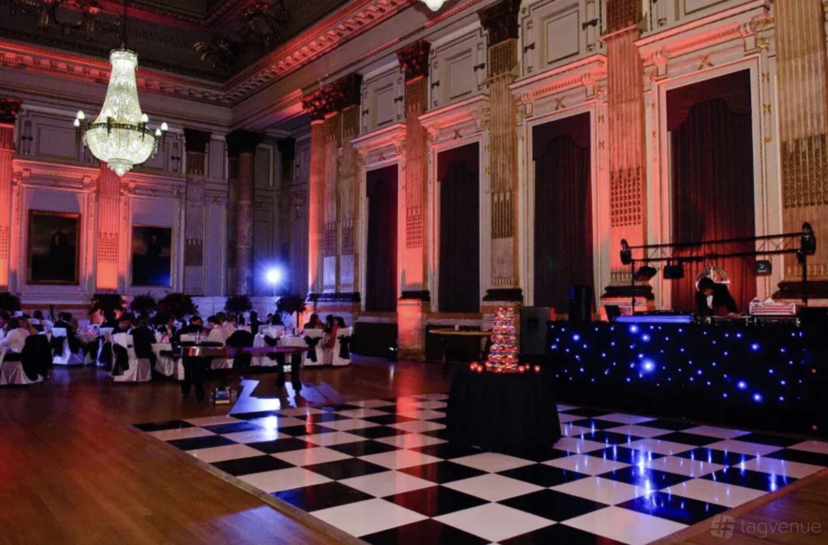 An event venue with a large chandelier, checkered dance floor, and uplighting at One Great George Street Great Hall.