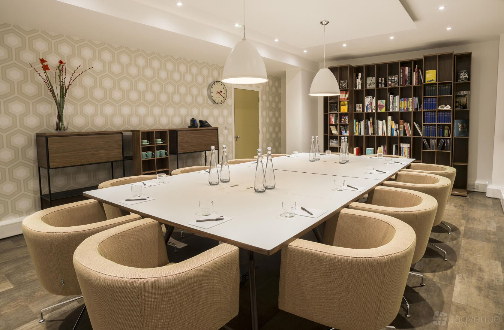 A meeting room with bookshelves, beige armchairs, pendant lights, and geometric wallpaper at The Derby.