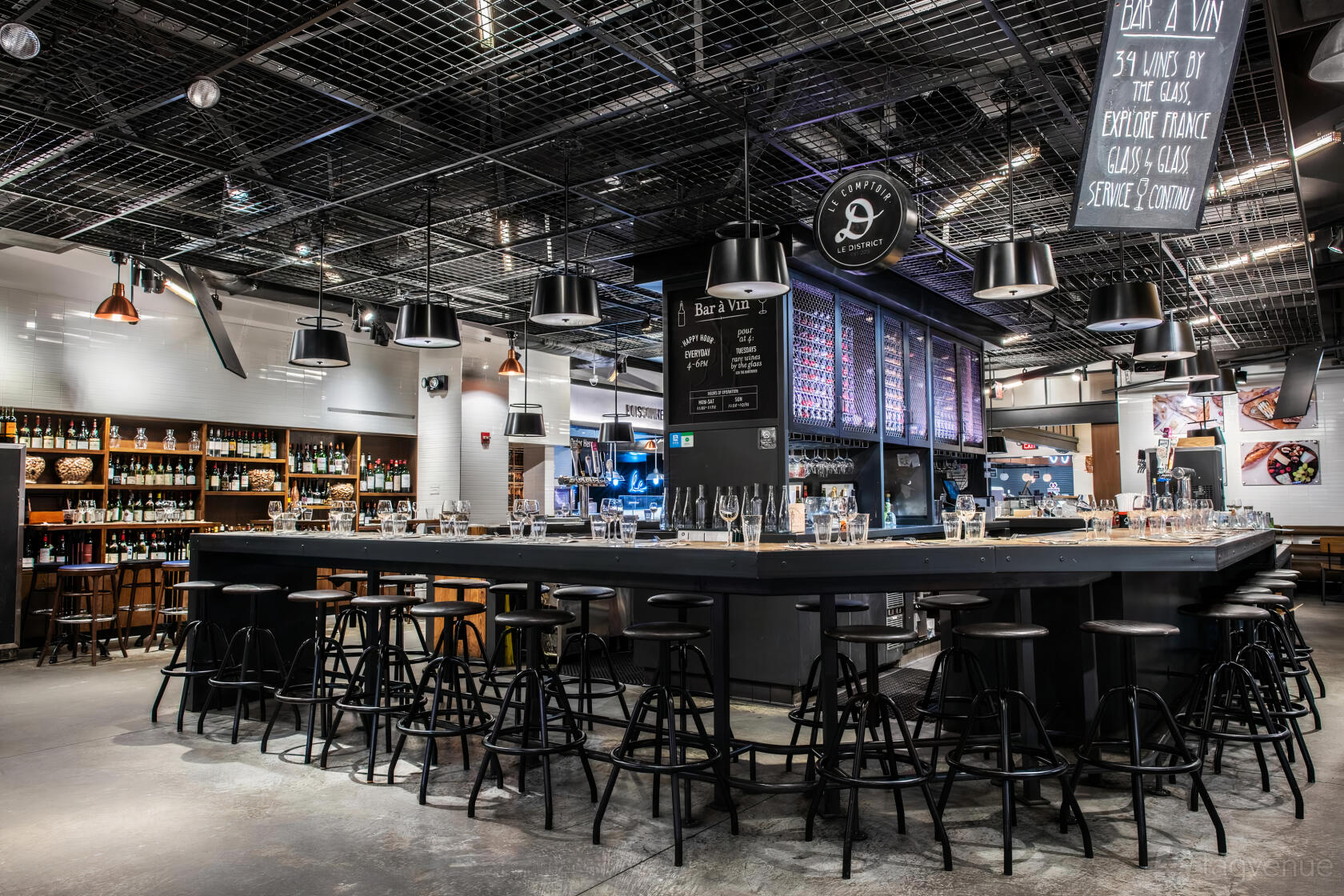 A bar in a restaurant with a large square counter, black barstools, glassware, and wine displays at Le District.