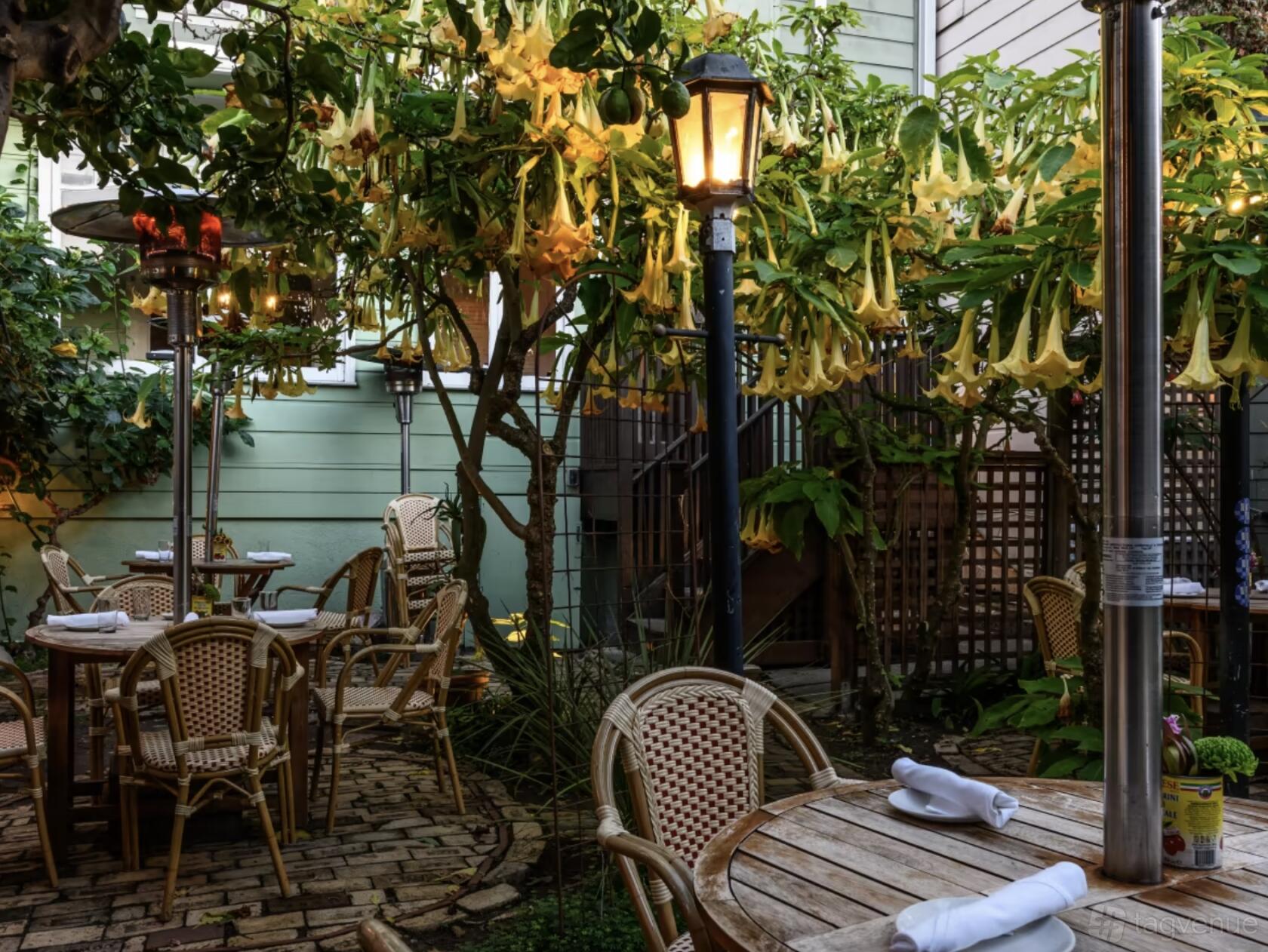 An outdoor restaurant space with woven bistro chairs, brick flooring, lush flowering trees, and lamplight at Blue Plate.