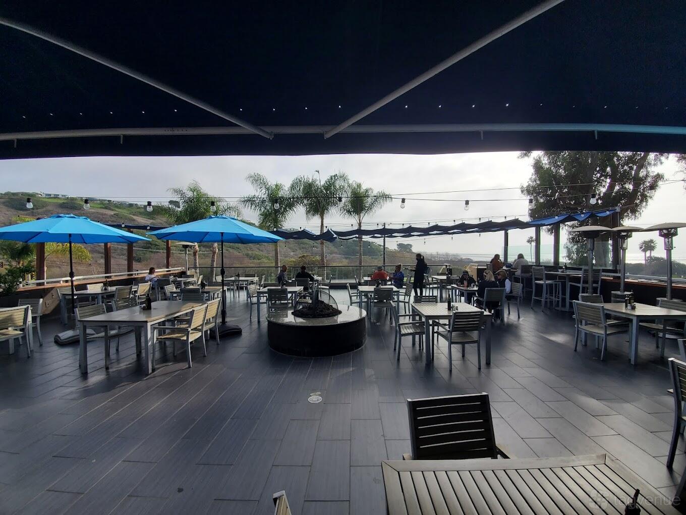 A restaurant terrace with ocean views, patio seating, blue umbrellas, and a central fire pit at Spruzzo Restaurant & Bar.