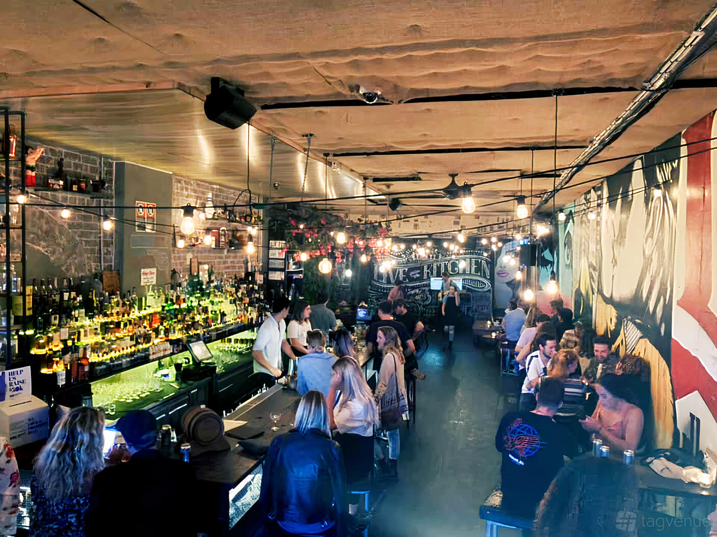 A cocktail bar with exposed brick walls, colorful wall murals, hanging bulbs, and a lively crowd at Pocket Bar.