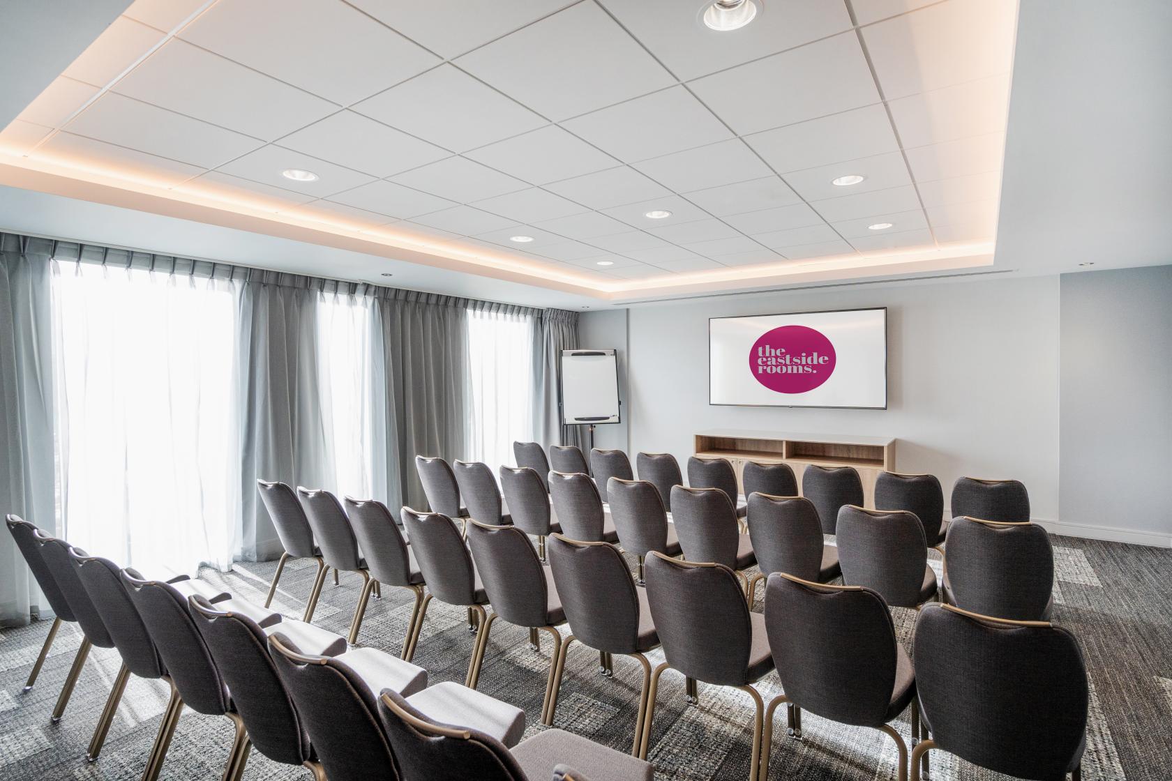 A meeting room with rows of gray chairs, a projector screen, and floor-to-ceiling windows at The Eastside Rooms.