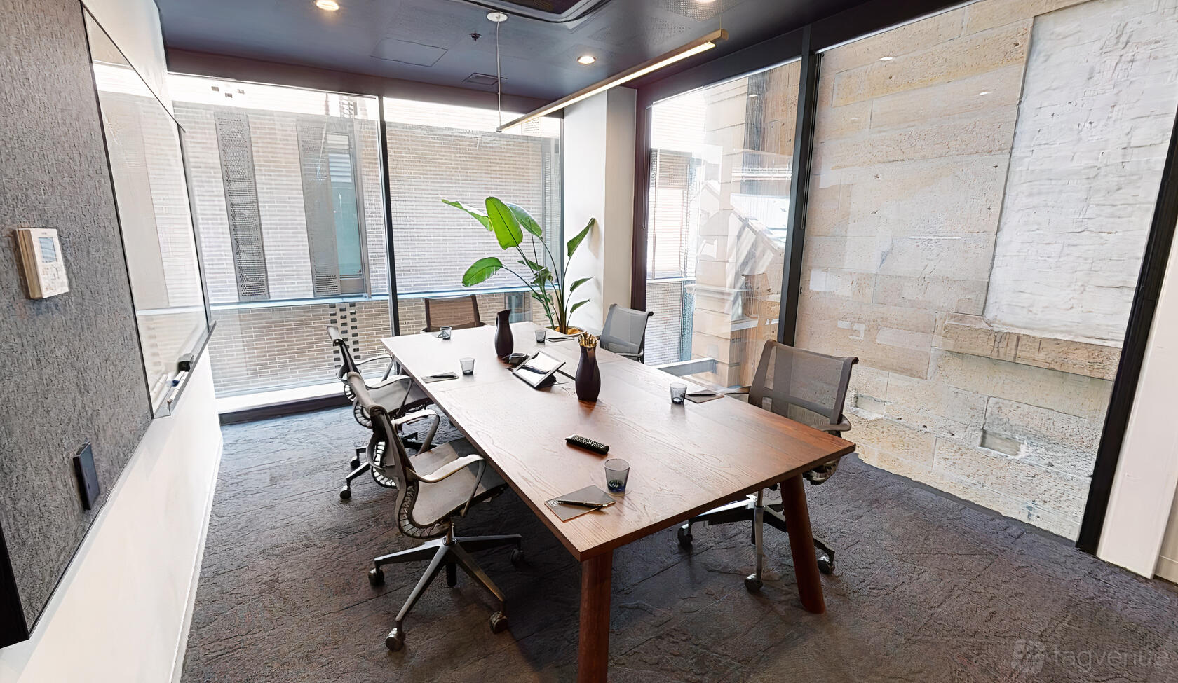 A coworking space with a wooden table, mesh office chairs, large windows, and a potted plant at Hub Custom House.