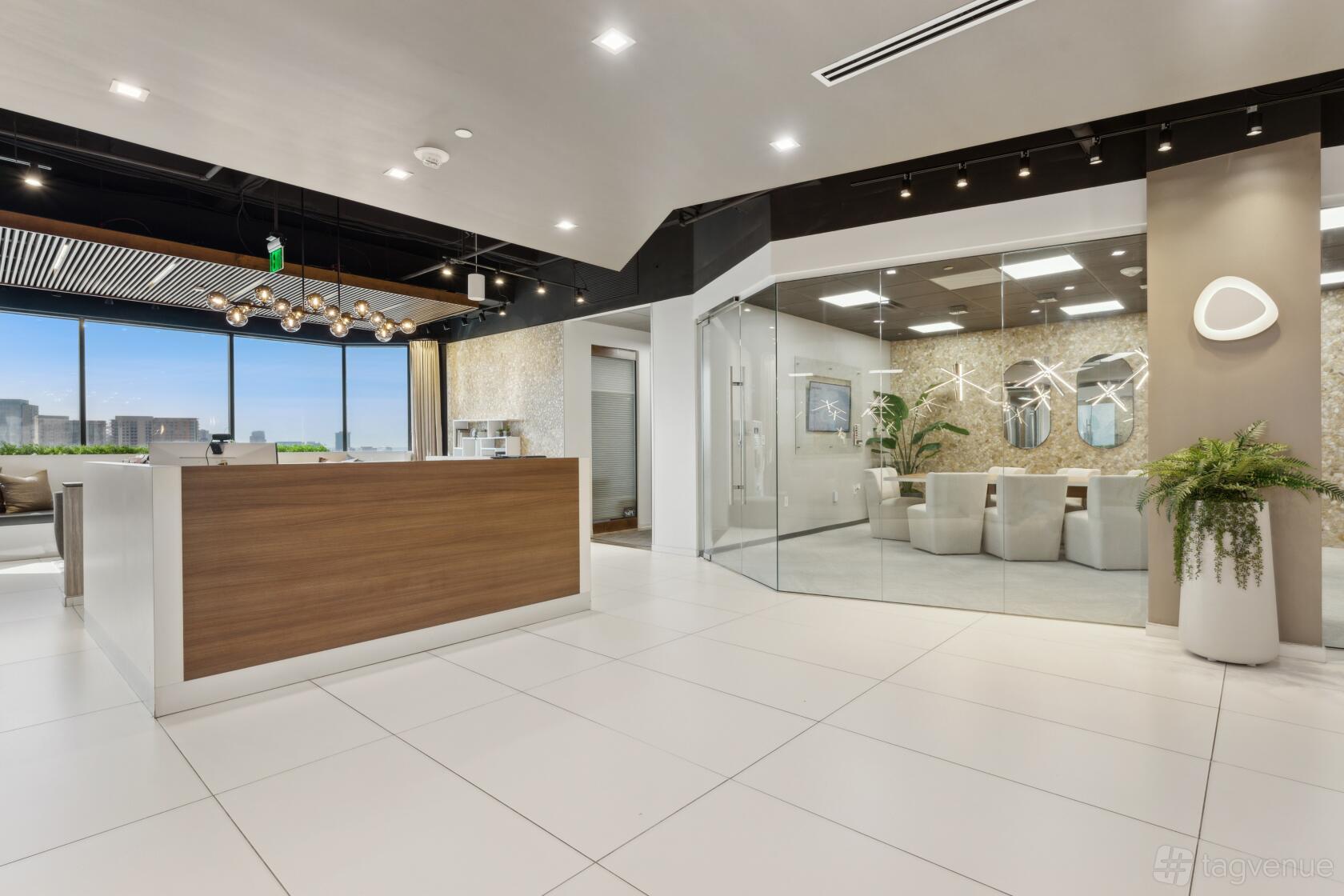A coworking reception area with a wooden front desk, glass-walled boardroom, white tile floor, and city views at Lucid Private Offices Dallas - Uptown.