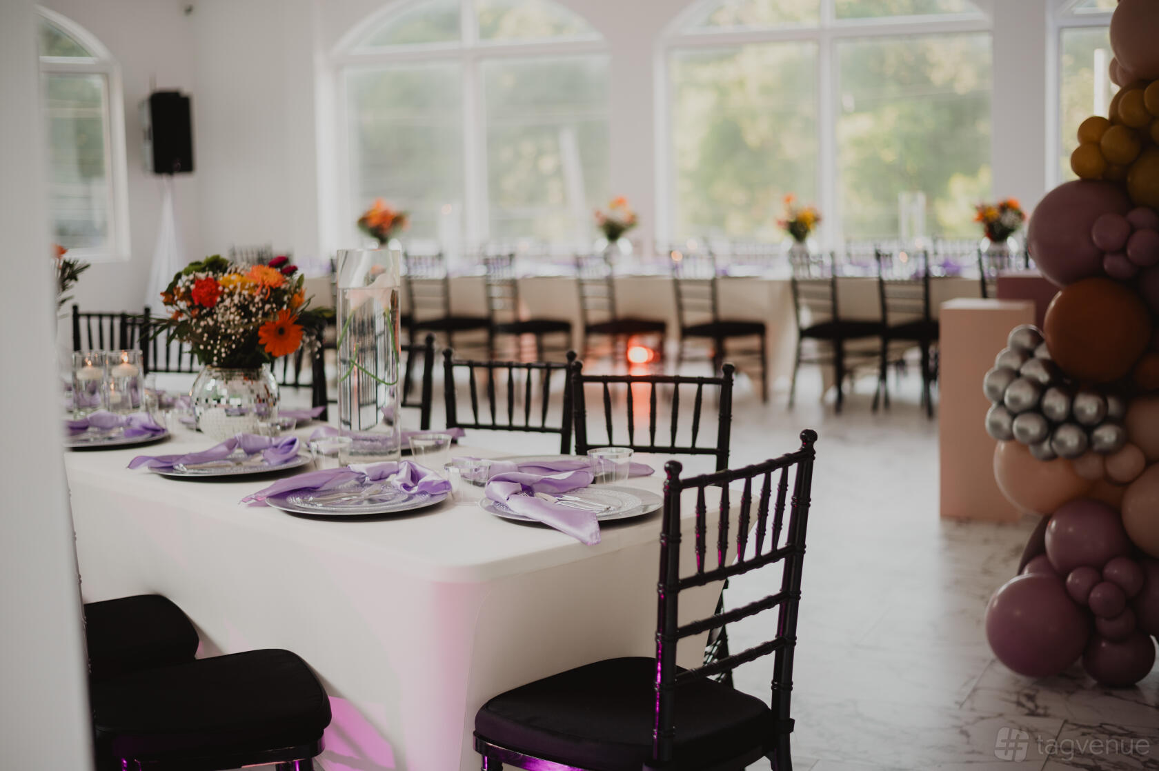 An event venue with white walls, arched windows, lilac napkins on banquet tables, and floral centerpieces at Luxx Lab Event Suite.