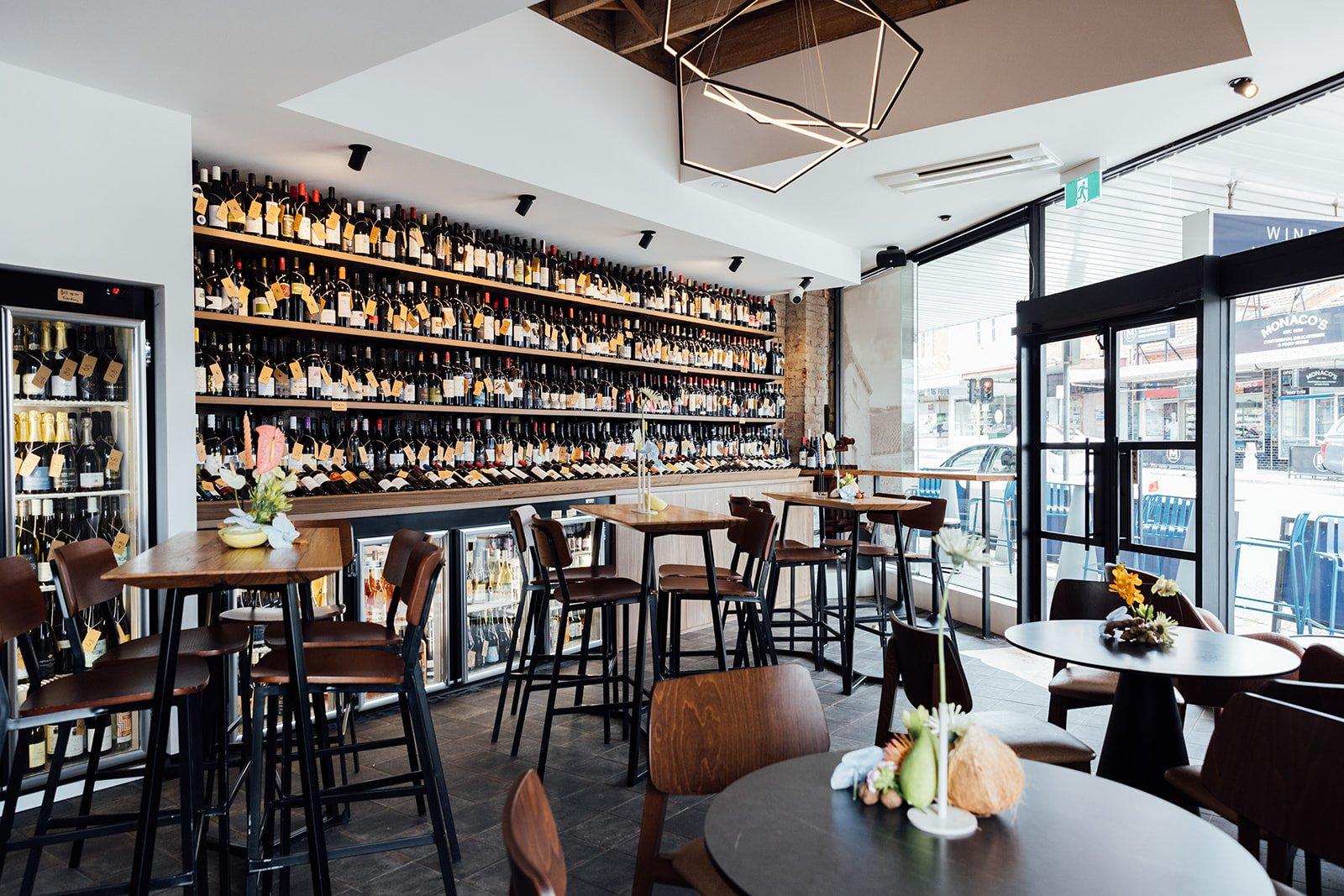A bar with floor-to-ceiling wine bottle displays, high tables, wood chairs, and pendant lighting at Running Postman Wine Bar.