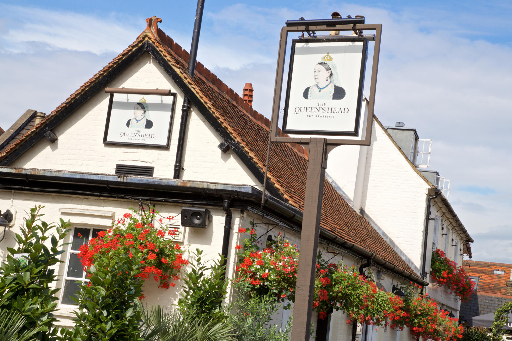 A white-walled pub with hanging red flower baskets and traditional signage at The Queen's Head Weybridge.