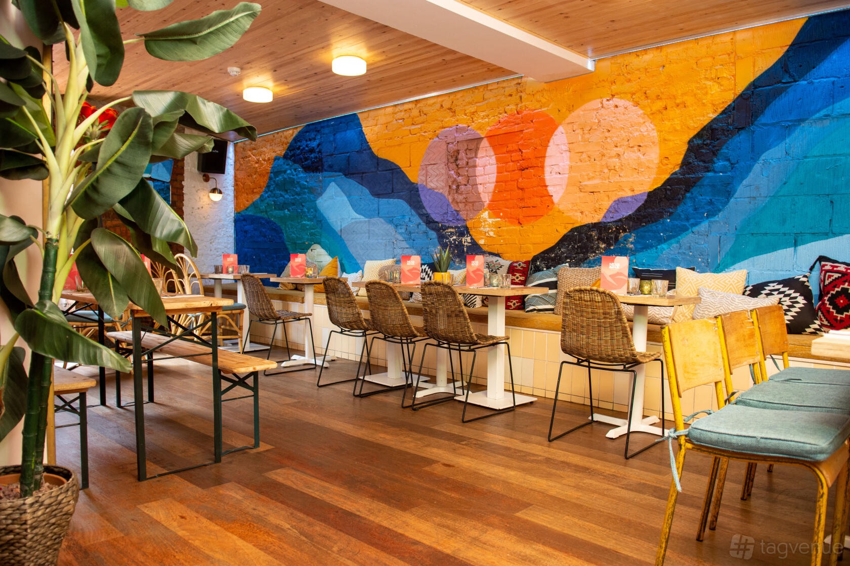 A function room with colorful mural walls, wooden flooring, and long tables with woven chairs at Studio48 Bar & Kitchen.