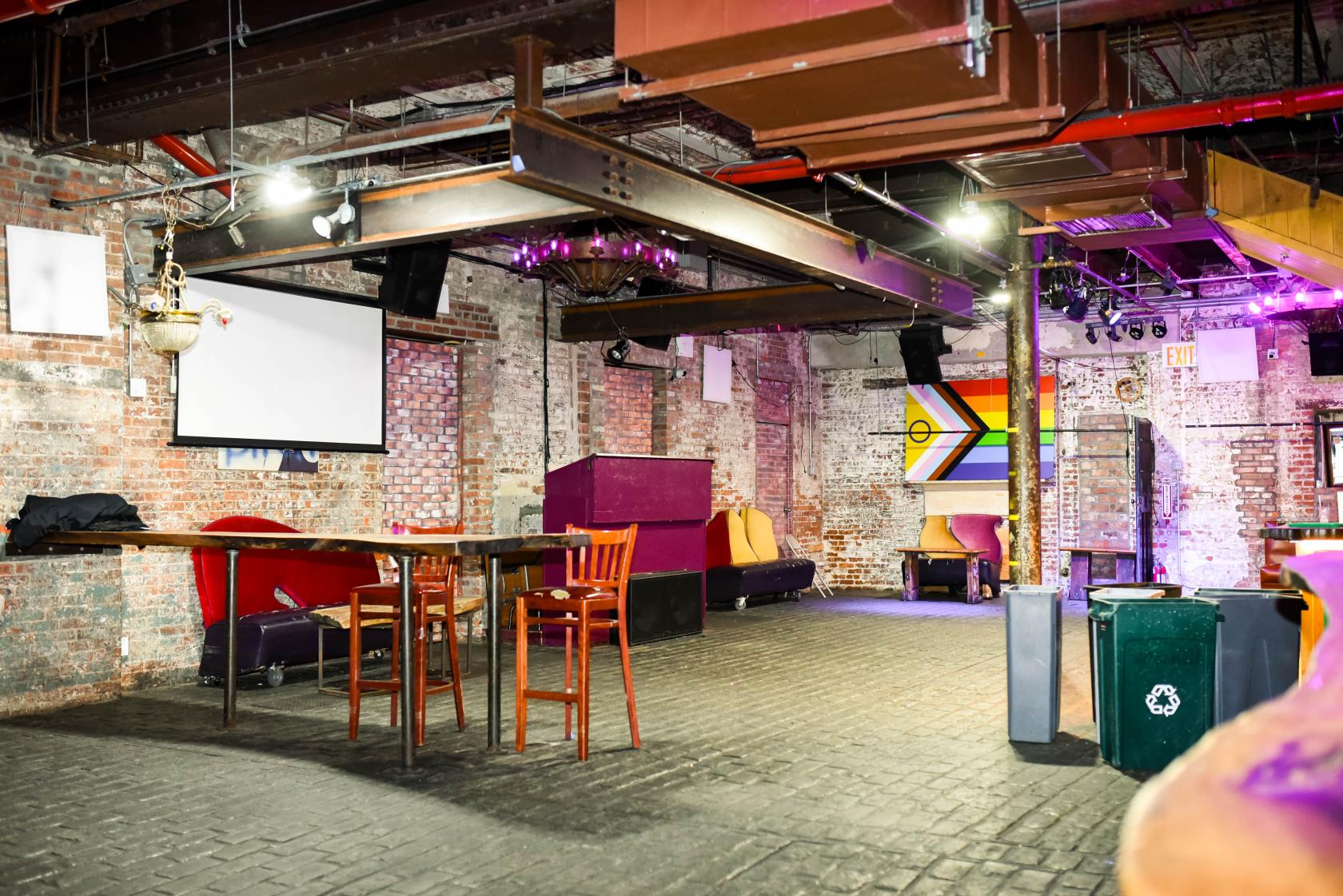 A nightclub dining room with exposed brick walls, industrial beams, stage lighting, and colorful seating at 3 Dollar Bill.