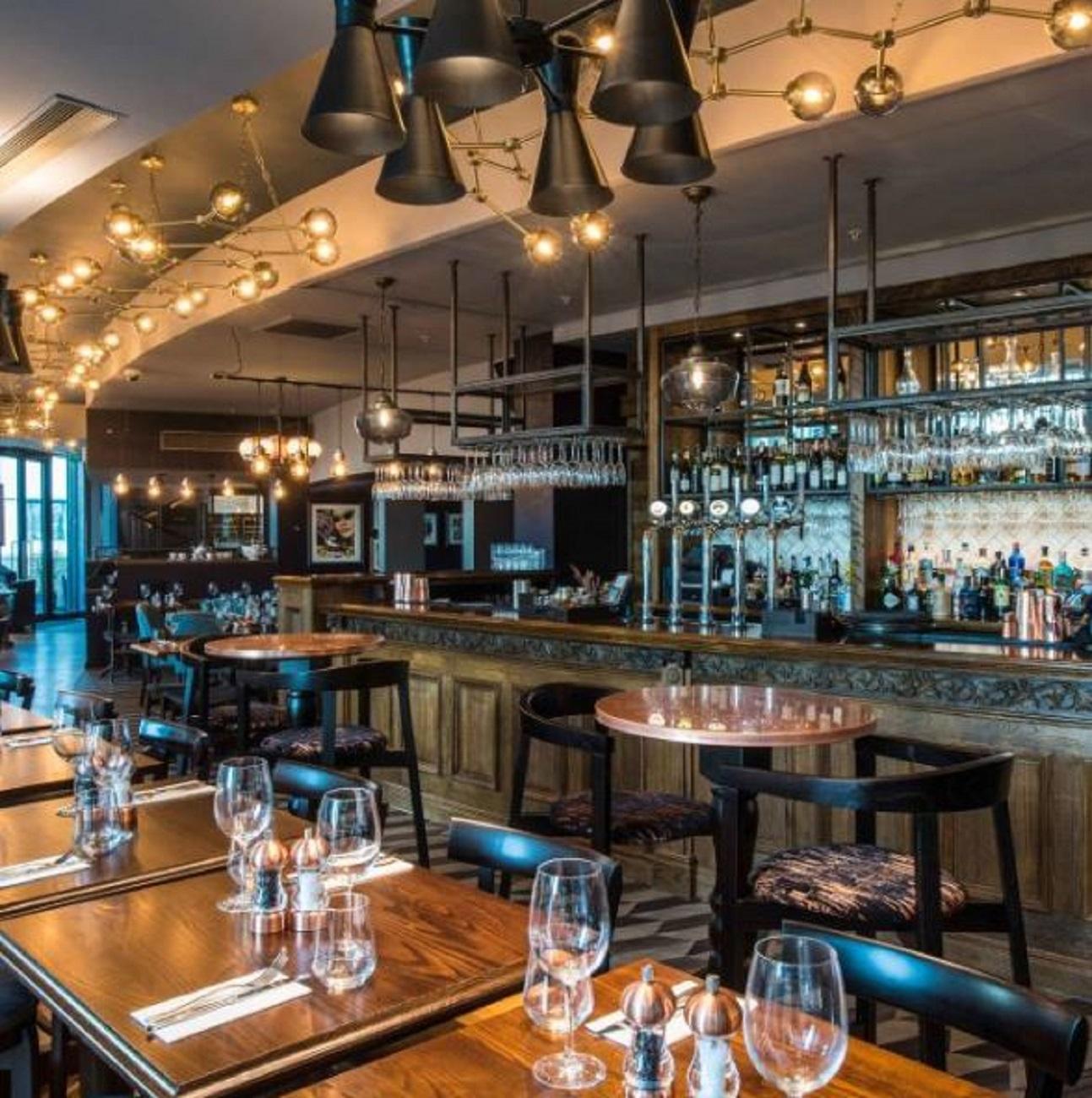 A restaurant with a long wooden bar, hanging pendant lights, and round tables set with glassware at Rocket.