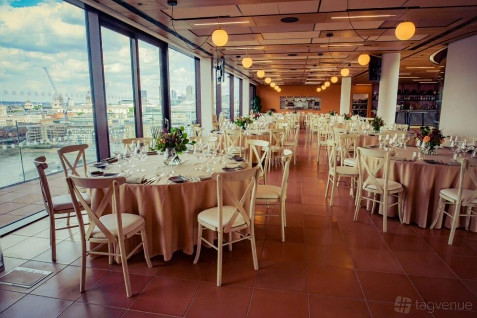 An event venue with floor-to-ceiling windows, round tables with beige linens, and city views at Rose Court Events.