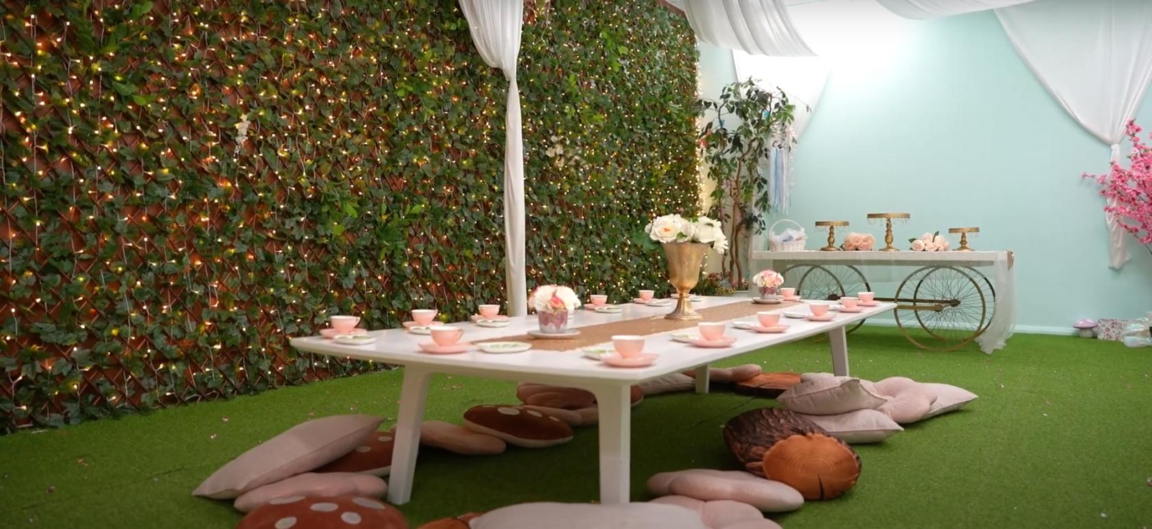 An event venue with a low table set for tea, decorative cushions, and a wall covered in greenery and string lights at The Enchanted Trove.